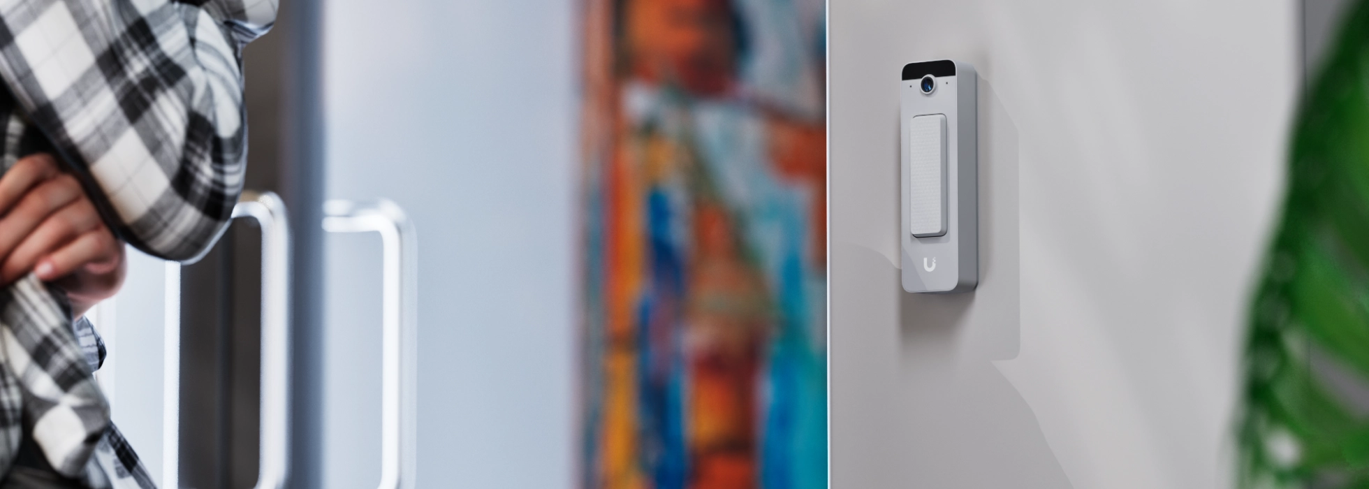 Ubiquiti Doorbell Lite zamontowane na białej ścianie, z widoczny tłem i lekką rozmytą barwą z kolorowymi obrazkami